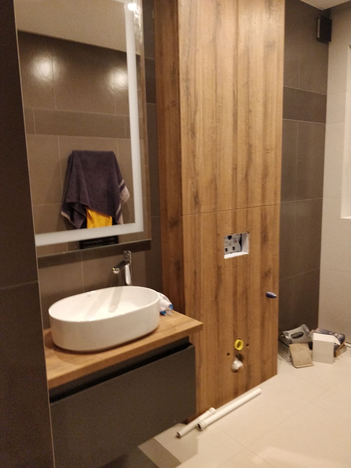 Nowoczesna łazienka w trakcie montażu: owalna umywalka nablatowa, drewniana szafka wisząca z otworem na stelaż podtynkowy, szare płytki na ścianach i podłodze, widoczne narzędzia i rury.