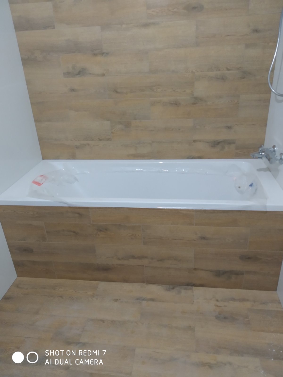 Nowa, biała wanna obudowana płytkami imitującymi drewno, z widoczną baterią i fragmentem słuchawki prysznicowej, zabezpieczona folią ochronną.