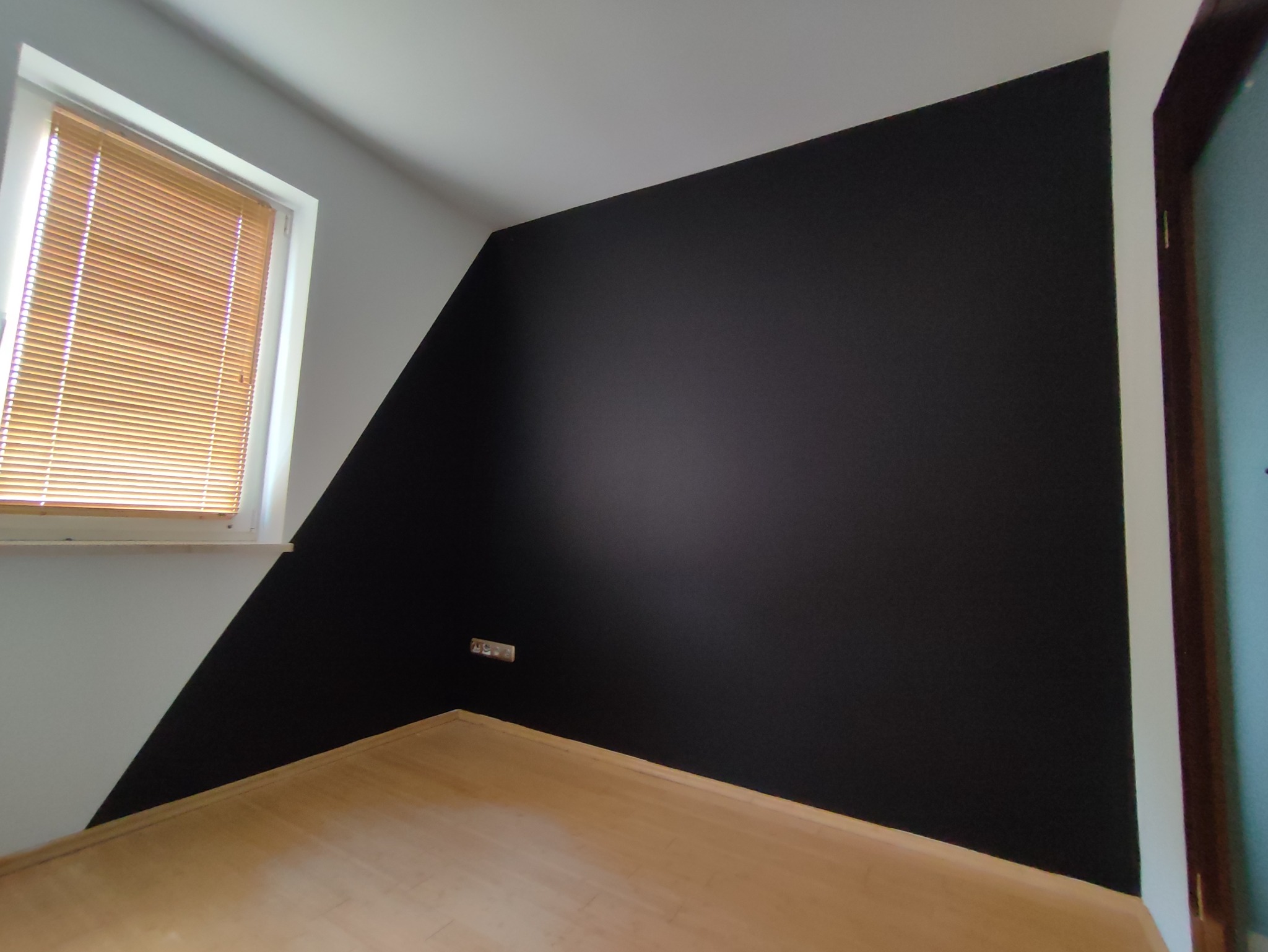 Ściana w pokoju pomalowana częściowo na czarno, z widocznym gniazdkiem elektrycznym i drewnianą podłogą. Okno z żaluzjami w tle.