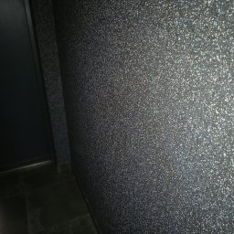 Usługi Remontowo-Budowlane WIETRZYŃSKI - Błyszcząca, ciemna ściana z efektem brokatu, widoczna w rogu pomieszczenia, z fragmentem ciemnych płytek podłogowych i drzwi po lewej stronie.