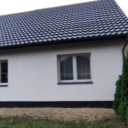 Usługi Remontowo-Budowlane WIETRZYŃSKI - Boczny widok otynkowanego, parterowego domu z ciemną dachówką i białymi oknami. W tle zieleń drzew i krzewów. Ujęcie z poziomu gruntu.