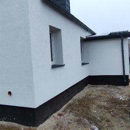 Usługi Remontowo-Budowlane WIETRZYŃSKI - Boczny widok otynkowanego domu jednorodzinnego z czarną podmurówką i białymi oknami. W tle widoczna osoba i przyczepka samochodowa.
