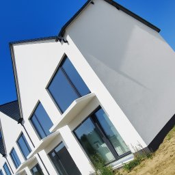 Usługi Remontowo-Budowlane WIETRZYŃSKI - Nowoczesny dom z białymi ścianami i dużymi oknami, widziany pod kątem z poziomu gruntu. Błękitne niebo w tle. Minimalistyczny design.
