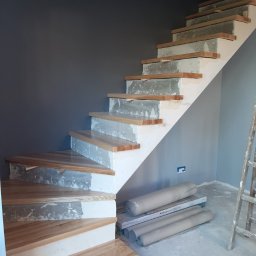 Usługi Remontowo-Budowlane WIETRZYŃSKI - Montaż drewnianych schodów z widocznymi elementami konstrukcyjnymi i przygotowaniem do wykończenia. W tle szara ściana i drabina.