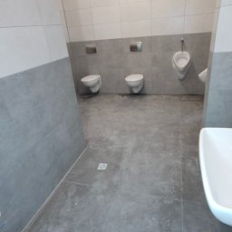 Usługi Remontowo-Budowlane WIETRZYŃSKI - Wykończona łazienka publiczna z szarymi płytkami, trzema toaletami i pisuarami. Widoczna umywalka z boku. Żółta instalacja pod sufitem. Minimalistyczny design.