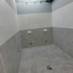 Usługi Remontowo-Budowlane WIETRZYŃSKI - Wykończona łazienka w szaro-białej kolorystyce z widocznymi przyłączami wodnymi i odpływem podłogowym. Minimalistyczny design, płytki ceramiczne na ścianach i podłodze.