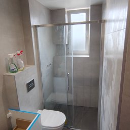 Usługi Remontowo-Budowlane WIETRZYŃSKI - Wykończona łazienka w szarościach z kabiną prysznicową typu walk-in, podwieszaną toaletą i częściowo zamontowaną szafką pod umywalkę. Widoczne środki czystości na półce.