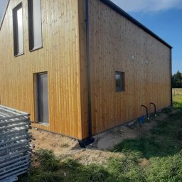 Usługi Remontowo-Budowlane WIETRZYŃSKI - Nowoczesny dom szkieletowy z drewnianą elewacją w pionowe deski, z widocznymi oknami i rynnami, na tle błękitnego nieba i zielonej trawy. Częściowo widoczne rusztowanie.