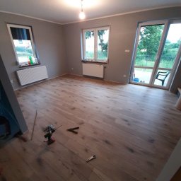 Usługi Remontowo-Budowlane WIETRZYŃSKI - Jasne panele podłogowe w szarym pokoju z oknami i narzędziami. Widoczne wkrętarka, kombinerki. Świeżo położona podłoga w nowym wnętrzu.