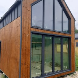 Usługi Remontowo-Budowlane WIETRZYŃSKI - Nowoczesny dom szkieletowy z drewnianą elewacją i dużymi oknami, widok z zewnątrz. Dom posadowiony na betonowych bloczkach, w tle zieleń.