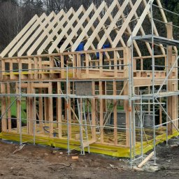 Usługi Remontowo-Budowlane WIETRZYŃSKI - Konstrukcja domu szkieletowego w trakcie budowy, widoczne drewniane ramy i rusztowania. Żółta folia izolacyjna na fundamencie. W tle drzewa i pochmurne niebo.