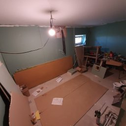 Usługi Remontowo-Budowlane WIETRZYŃSKI - Wnętrze w trakcie remontu: płyty karton-gips na ścianach, podłoga częściowo zakryta kartonem, widoczne narzędzia i materiały budowlane. Żarówka oświetla pomieszczenie.