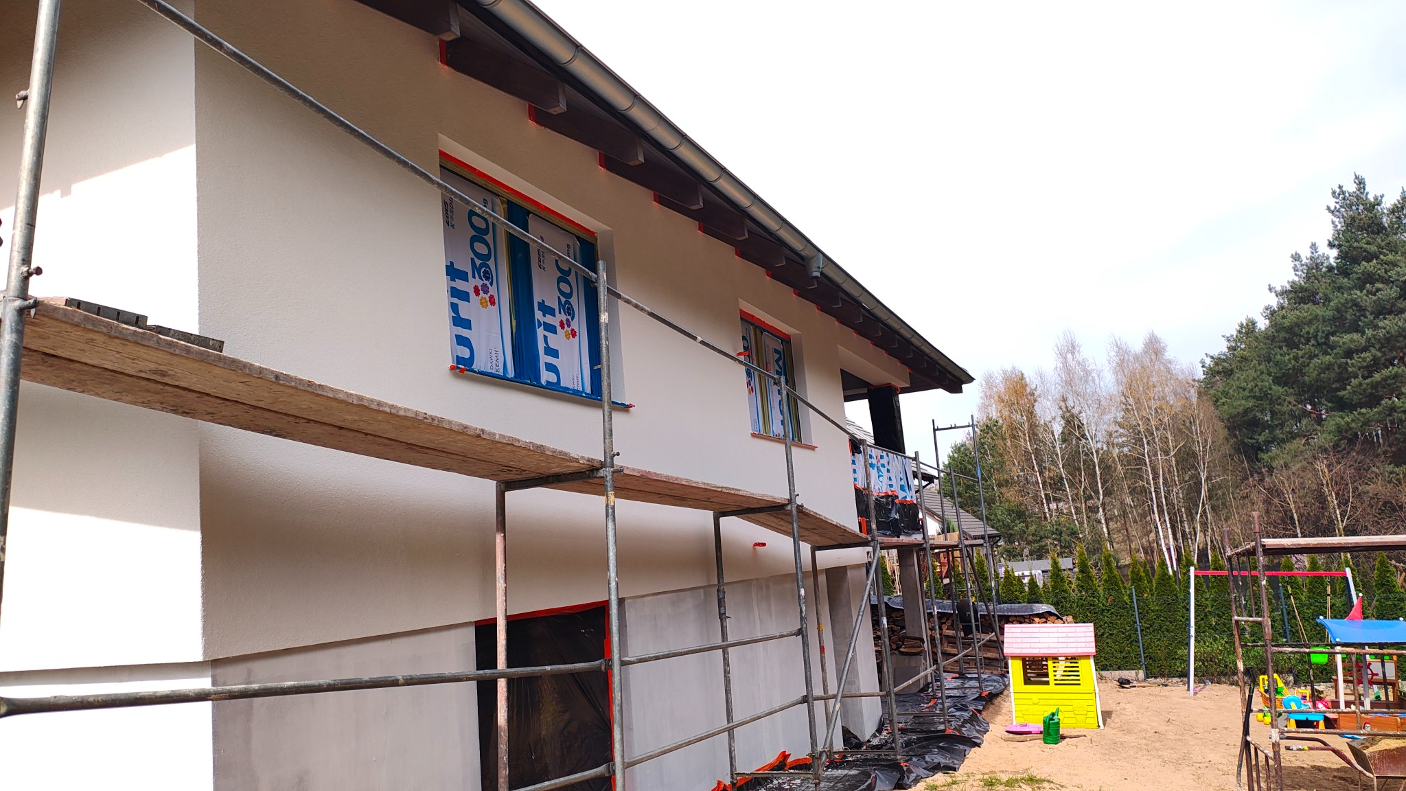 Ocieplony, biały dom jednorodzinny w trakcie prac wykończeniowych. Widoczne rusztowania, zabezpieczone okna i fragment placu zabaw w tle.