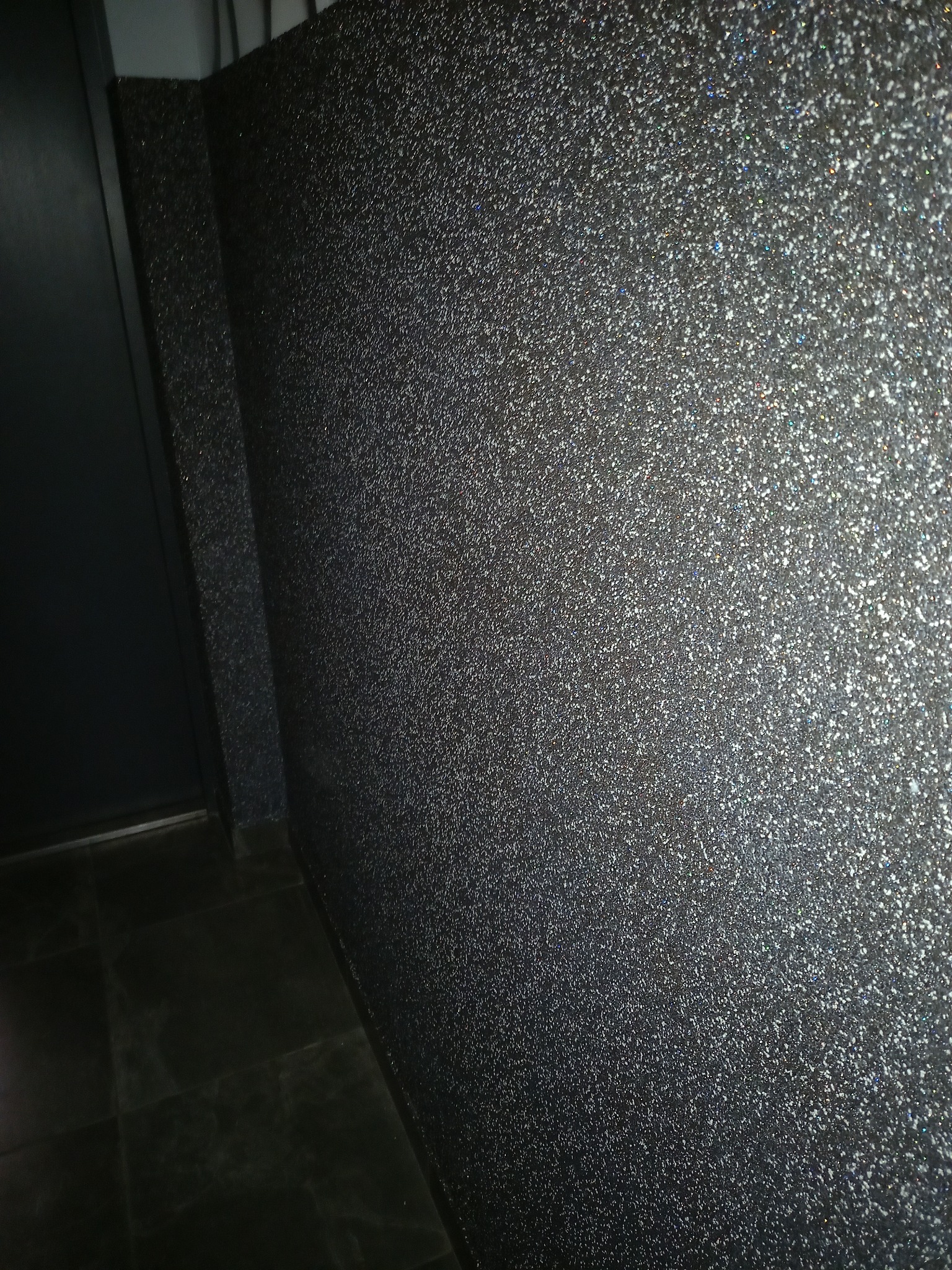 Błyszcząca, ciemna ściana z efektem brokatu, widoczna w rogu pomieszczenia, z fragmentem ciemnych płytek podłogowych i drzwi po lewej stronie.