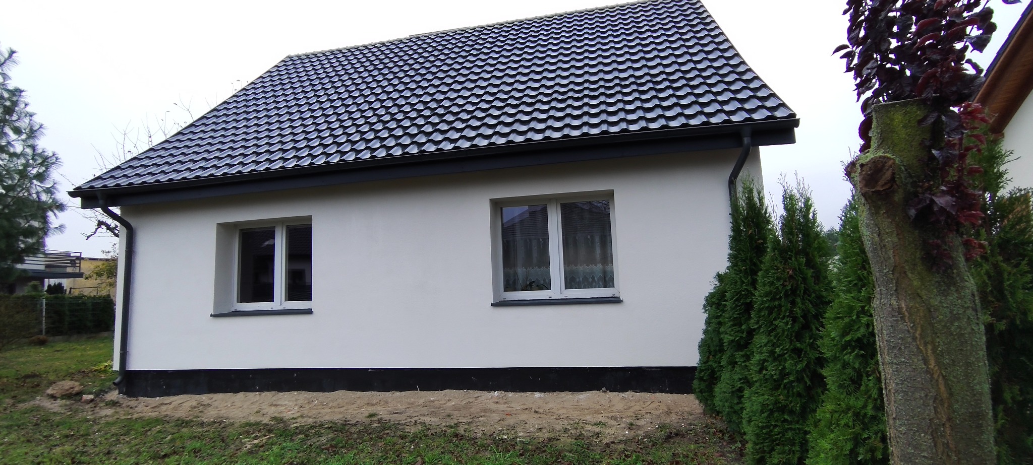 Boczny widok otynkowanego, parterowego domu z ciemną dachówką i białymi oknami. W tle zieleń drzew i krzewów. Ujęcie z poziomu gruntu.