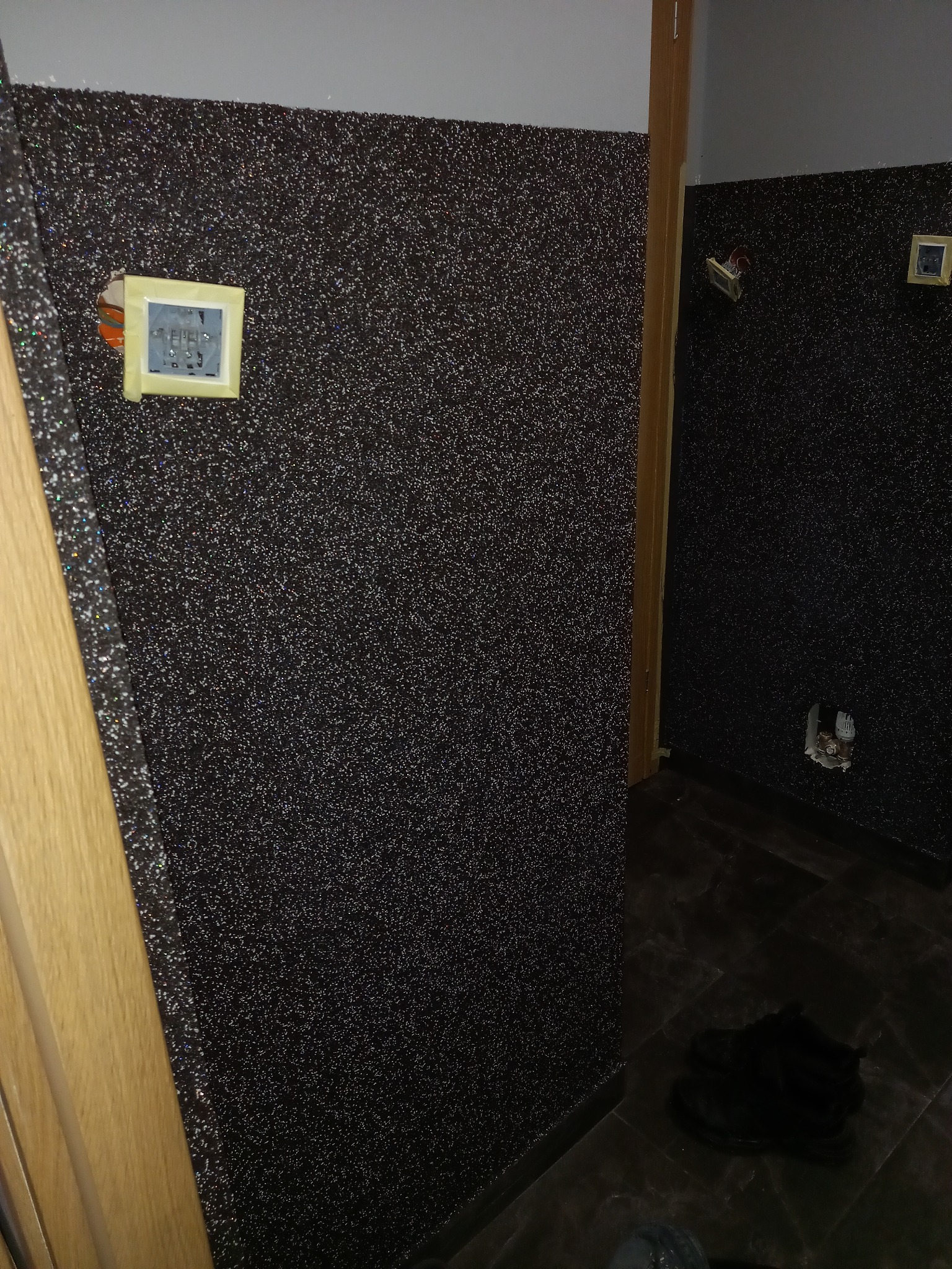 Ściana w korytarzu pokryta ciemną, błyszczącą tapetą strukturalną. Widoczne gniazdka elektryczne w trakcie montażu i fragment drewnianej futryny drzwiowej.