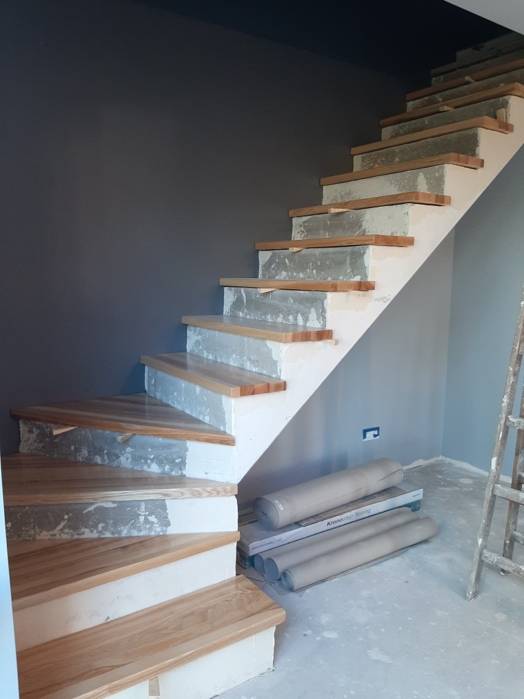 Montaż drewnianych schodów z widocznymi elementami konstrukcyjnymi i przygotowaniem do wykończenia. W tle szara ściana i drabina.