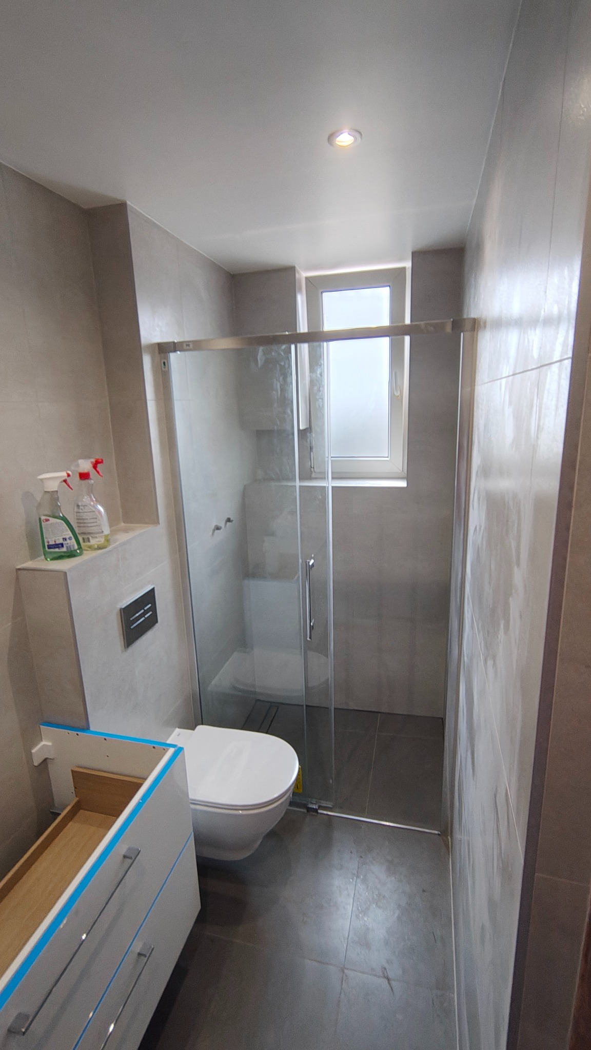 Wykończona łazienka w szarościach z kabiną prysznicową typu walk-in, podwieszaną toaletą i częściowo zamontowaną szafką pod umywalkę. Widoczne środki czystości na półce.