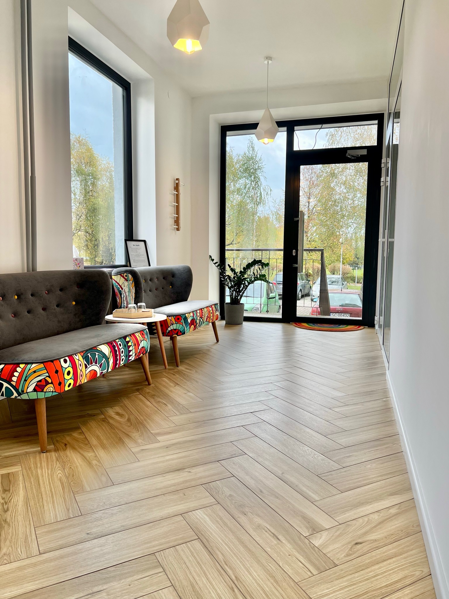 Jasne wnętrze poczekalni z drewnianą podłogą ułożoną w jodełkę, szarymi sofami z kolorowymi wzorami, białym stolikiem i rośliną doniczkową przy przeszklonych drzwiach wejściowych.