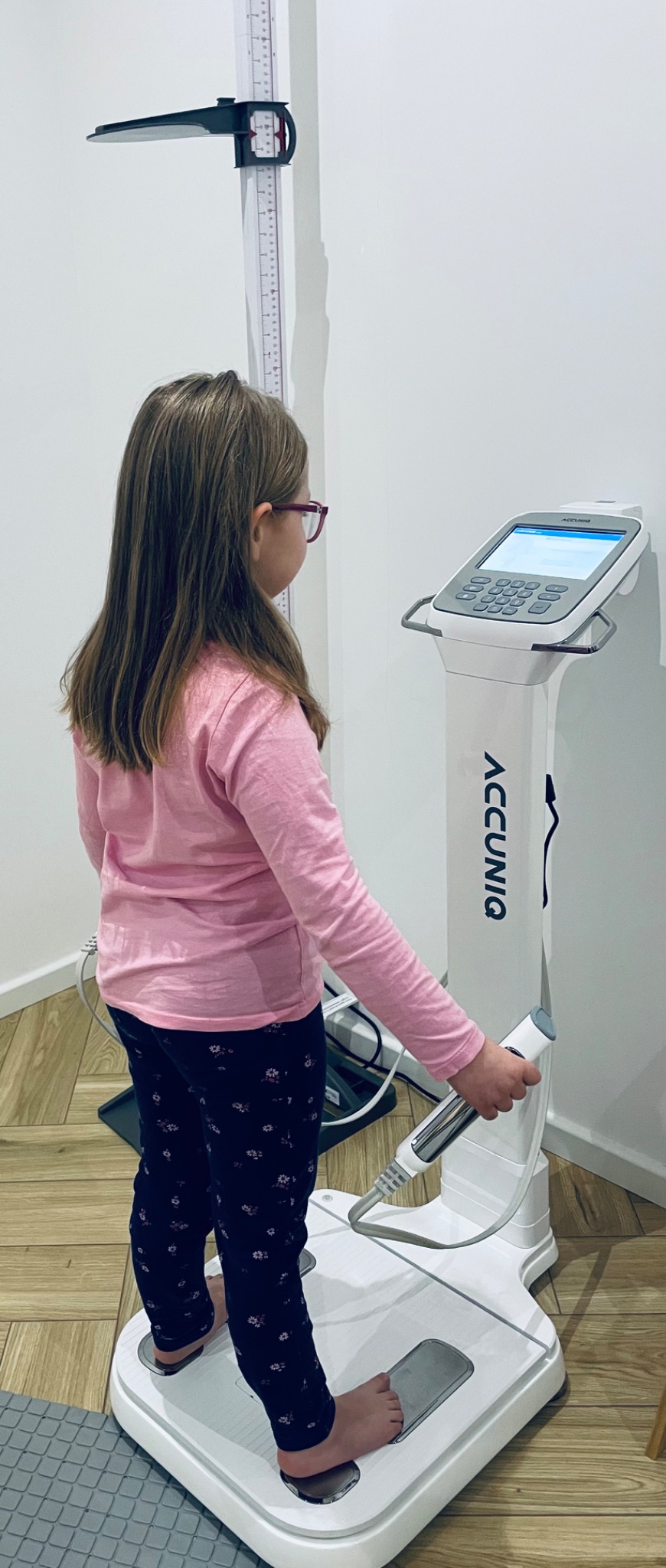 Dziewczynka w okularach stoi na analizatorze składu ciała, trzymając uchwyt, obok stoi wzrostomierz.
