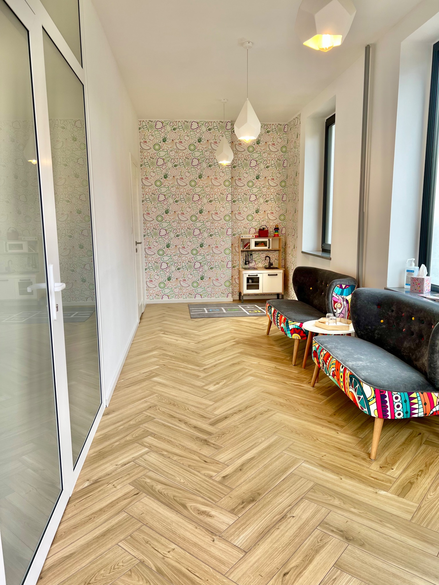 Jasne wnętrze poczekalni z wzorzystą tapetą w owoce, dwoma szarymi sofami z kolorowymi bokami, drewnianą podłogą w jodełkę i dziecięcą kuchnią zabawkową w tle.