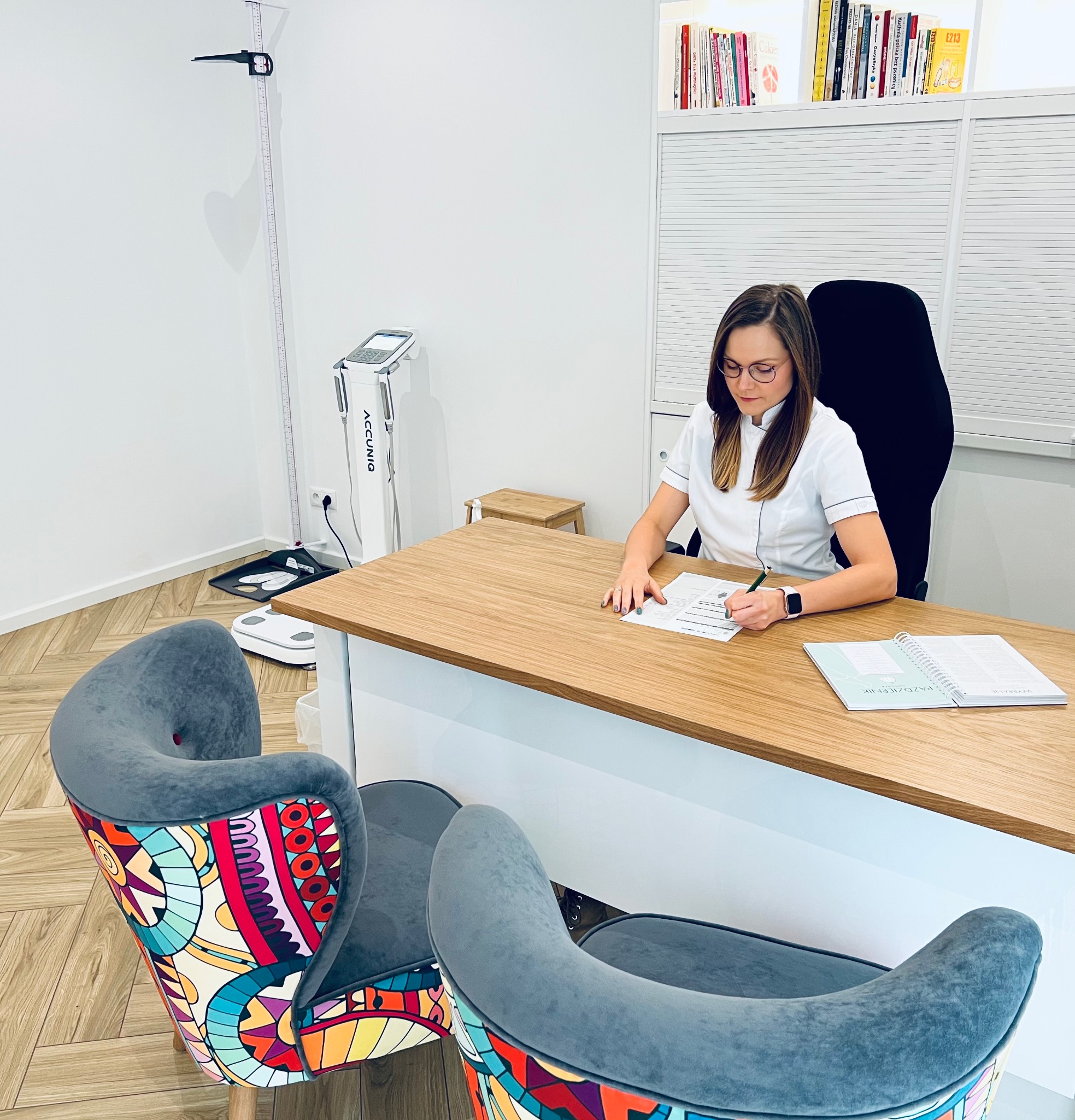 Kobieta w białym uniformie, z zegarkiem na ręku, wypełnia dokumenty przy biurku w gabinecie, obok waga analityczna i kolorowe krzesła.