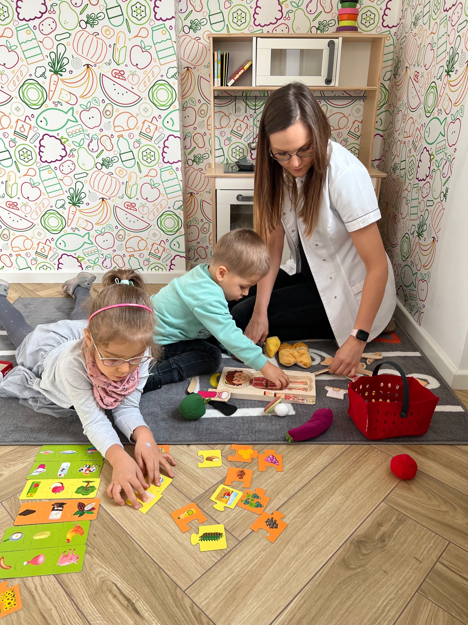 Dietetyk w białym fartuchu pomaga dzieciom układać puzzle z jedzeniem w gabinecie z tapetą w owoce i warzywa.