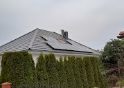 Panele słoneczne na szarym dachu domu jednorodzinnego, widoczne zza rzędu wysokich, zielonych tuj. Pochmurne niebo w tle.