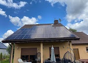 Panele słoneczne na dachu domu jednorodzinnego, widoczne odbicia chmur na powierzchni paneli, brązowa dachówka, jasna elewacja budynku, błękitne niebo.