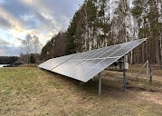 Instalacja paneli słonecznych na łące, widok z boku, w tle jezioro i las, pochmurne niebo.