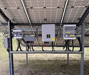 Widok od spodu na panele słoneczne z widocznymi inwerterami i skrzynkami przyłączeniowymi zamontowanymi na metalowej konstrukcji. Kabelki i przewody starannie poukładane.