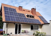 Instalacja fotowoltaiczna na dachu domu jednorodzinnego z czerwoną dachówką, widoczne dwa kominy, okna dachowe i panele umieszczone na kilku płaszczyznach dachu, zieleń w tle.