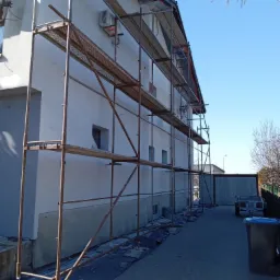 Dom jednorodzinny w trakcie prac elewacyjnych, widoczne rusztowanie, świeżo otynkowana ściana w jasnym kolorze, fragment dachu z elementami zabezpieczającymi, klimatyzatory na ścianie, błękitne...