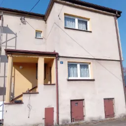 Dwukondygnacyjny dom z beżową elewacją w trakcie ocieplania, widoczne rusztowanie i fragment ocieplenia styropianem przy balkonie, w tle błękitne niebo.