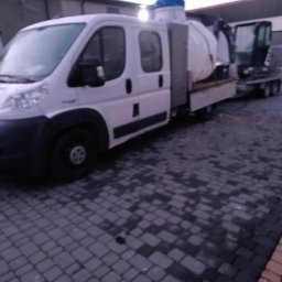 Firma Warte Jacek Cywiński - Specjalistyczny pojazd z cysterną i mini koparką na przyczepie, prawdopodobnie do oczyszczania kanalizacji, zaparkowany na brukowanym placu.