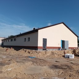 Firma Warte Jacek Cywiński - Budynek po ociepleniu elewacji z widocznymi otwartymi drzwiami i przygotowanym terenem wokół. Widoczne materiały budowlane i sprzęt. Jasne niebo.