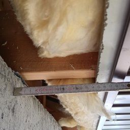 Firma Warte Jacek Cywiński - Pomiar grubości izolacji z wełny mineralnej w konstrukcji dachu za pomocą rozłożonej miarki. Widoczne elementy drewniane i metalowe konstrukcji.