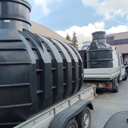 Firma Warte Jacek Cywiński - Czarne zbiorniki na szarych przyczepach transportowane na posesję z kostki brukowej, w tle budynek z panelami słonecznymi i błękitne niebo z chmurami.