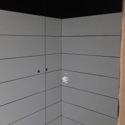 Firma Warte Jacek Cywiński - Wykończona łazienka z białymi, poziomymi płytkami i czarnym pasem u góry. Widoczny odpływ liniowy w podłodze i dwa punkty instalacyjne na ścianie.