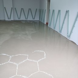 Firma Warte Jacek Cywiński - Wylany, jasny podkład podłogowy w pomieszczeniu z białymi ścianami. Na ścianach widoczne pionowe, metalowe listwy. Na podłodze wzór w kształcie plastra miodu.