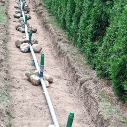 Firma Warte Jacek Cywiński - Instalacja drenażowa z rur PVC i kamieni w wykopie, obok żywopłotu. Widok z góry, ukazujący etap prac przy systemie odprowadzania wody.