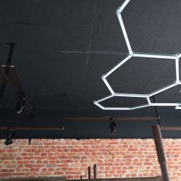 Firma Warte Jacek Cywiński - Czarne wnętrze z ceglaną ścianą, oświetlone lampami w kształcie plastra miodu i reflektorami. Widoczne drewniane belki i sufit z widoczną fakturą.