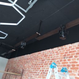 Firma Warte Jacek Cywiński - Montaż oświetlenia w biurze w Kole: czarny sufit z lampami na drewnianych belkach, ceglana ściana i drabina. Prace wykończeniowe w toku.