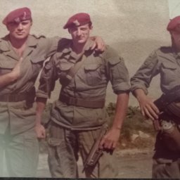 Firma Warte Jacek Cywiński - Trzech żołnierzy w mundurach i bordowych beretach, jeden z karabinem AK-47, drugi z pistoletem, pozują na tle jasnej ściany. Zdjęcie ma ciepłą tonację i wygląda na stare.