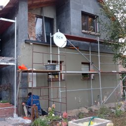 Firma Warte Jacek Cywiński - Dom w trakcie ocieplania: szare płyty izolacyjne na elewacji, rusztowania, pracownik w niebieskiej koszuli. Widoczna antena satelitarna i fragment ogrodu.