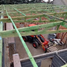 Konstrukcja drewniana dachu w trakcie budowy, widoczny miniładowarka Kingway i bloczki fundamentowe, w tle drzewa iglaste.