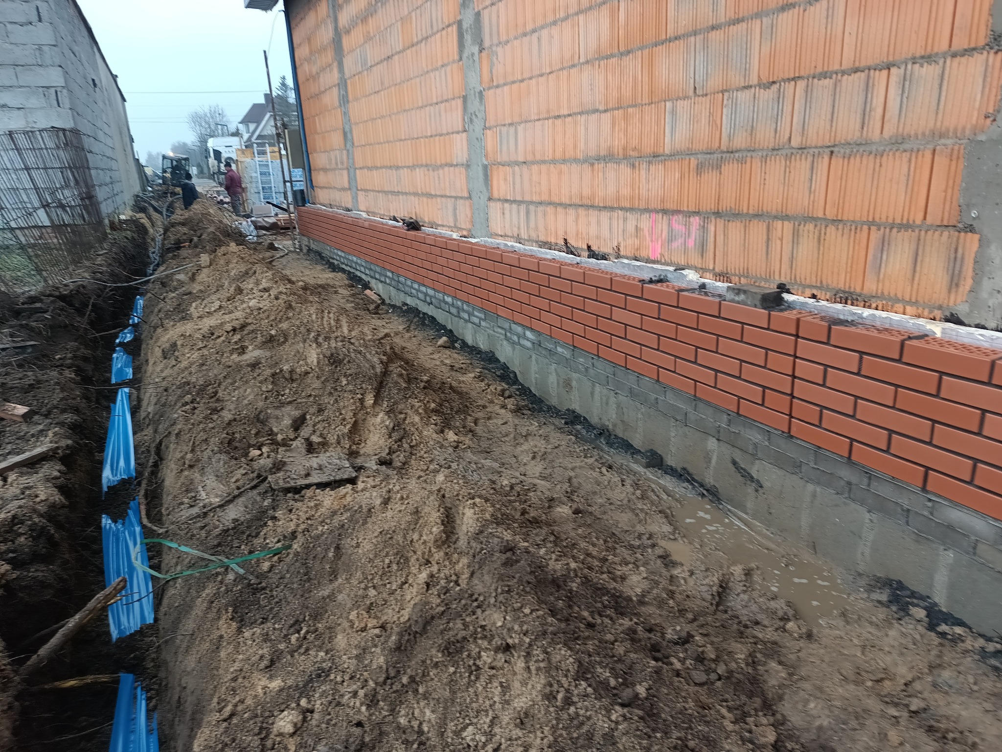 Murowanie elewacji z cegły klinkierowej na fundamencie. Wykop pod instalacje sanitarne z niebieskimi rurami w ziemi. Prace budowlane w toku, widoczni pracownicy w tle.