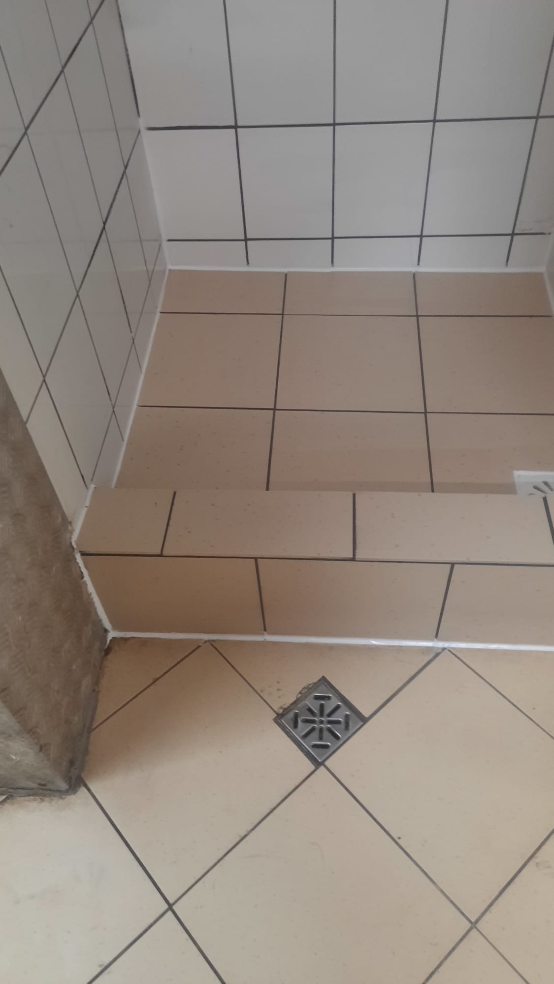 Wykończona kabina prysznicowa z beżowymi płytkami, widoczny odpływ liniowy i biała fuga. Płytki ułożone w dwóch różnych wzorach, na ścianach prosto, na podłodze w karo.