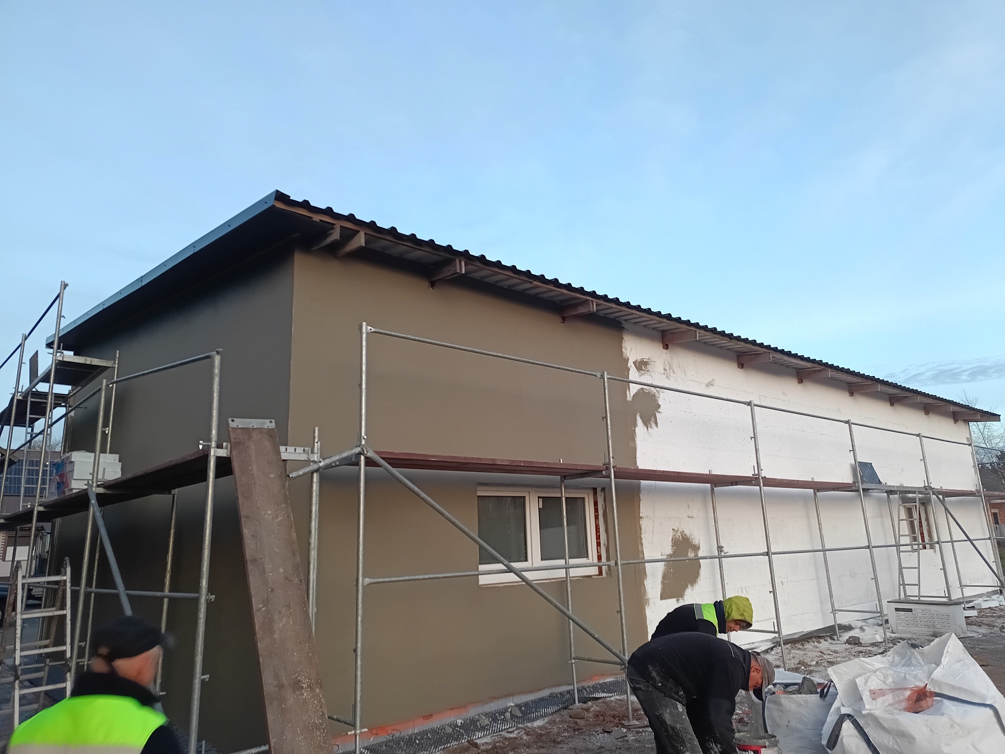 Budynek w trakcie ocieplania elewacji: część ściany z ociepleniem, część z nałożonym tynkiem. Widoczne rusztowania i pracownicy w trakcie pracy.