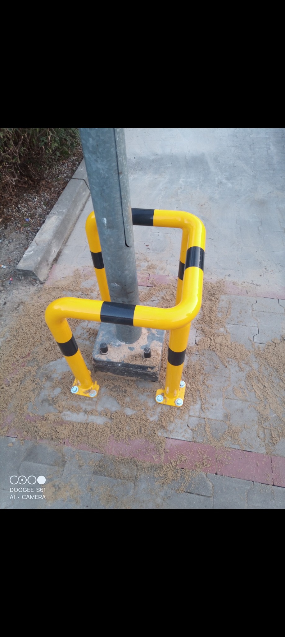 Żółto-czarna bariera ochronna wokół metalowego słupka, zabezpieczona śrubami na kostce brukowej. Widoczne ślady piasku.